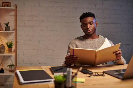 young thoughtful african american freelancer looking at notebook while working at home at nightの写真素材