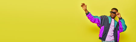 thrilled african american man in sunglasses and bright jacket waving hand on yellow backdrop, banner