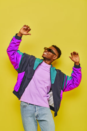 happy african american man in stylish casual attire and sunglasses dancing on yellow backdrop