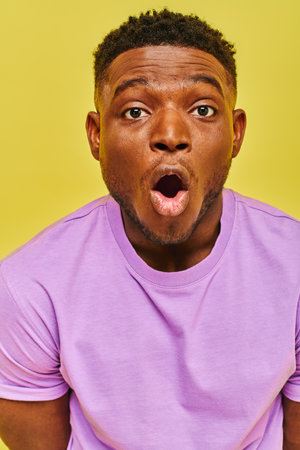 amazed african american man in purple t-shirt with open mouth looking at camera on yellow backdrop