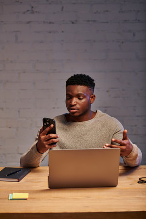 serious african american freelancer with smartphone gesturing during video chat on laptop at homeの写真素材