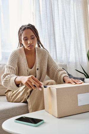 young attractive african american woman in homewear using stationery knife to open her parcel