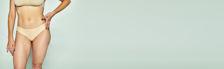 cropped shot of young woman standing in beige lingerie with hand on hip on grey background, bannerの写真素材