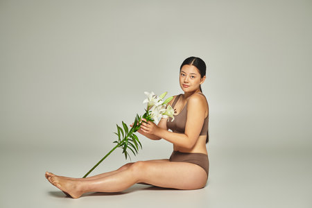 young and brunette asian woman in underwear holding flowers and sitting on grey background, liliesの写真素材