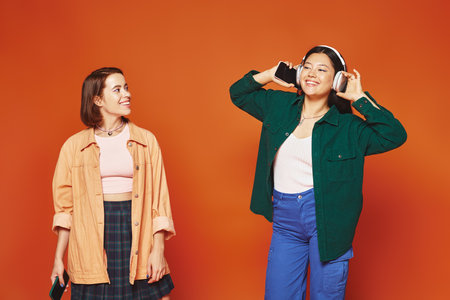 happy woman with smartphone looking at her asian friend in headphones on orange backgroundの写真素材