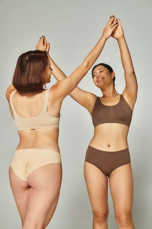 young happy interracial women in lingerie high-fiving each other on grey background, body positiveの写真素材