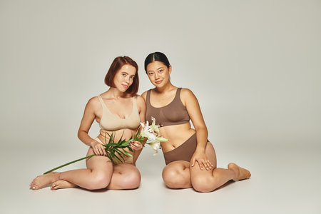 young multicultural women in underwear sitting together with flowers on grey background, liliesの写真素材
