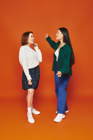 joyful multicultural female friends in vibrant attire looking at each other on orange backgroundの写真素材