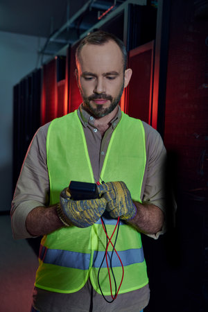 attractive bearded man in safety vest and gloves looking at detector in his hands, data centerの写真素材