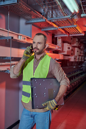 concentrated technician in safety vest and gloves talking by phone and looking away during work