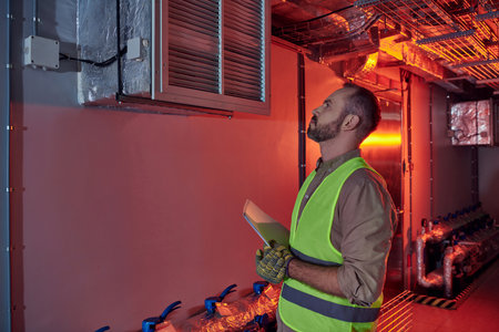 attractive concentrated technician in safety gloves and vest holding papers and looking up, dataの写真素材