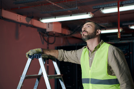 handsome pensive technician in safety vest standing next to step ladder and looking up, data centerの写真素材