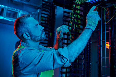 good looking concentrated technician in safety gloves and vest inspecting cables, data center