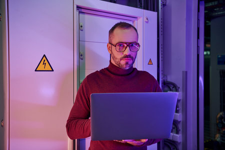 good looking dedicated specialist with glasses and beard holding his laptop and looking at camera