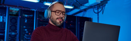 joyous specialist in turtleneck with headphones working hard on his laptop, data center, banner