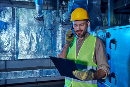 jolly hardworking technician in safety helmet and clothes looking at clipboard and talking by phoneの写真素材
