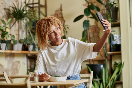 cheerful african american girl in braces taking selfie while eating fresh vegan salad in cafeの写真素材