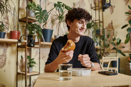 happy and young curly man holding tofu burger and looking at smartphone on table in vegan cafeの写真素材