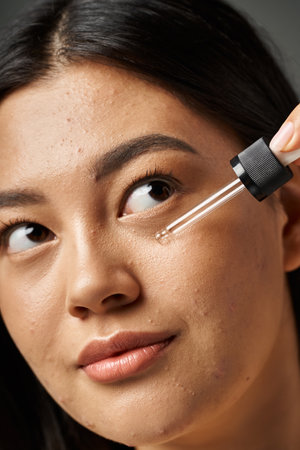 young woman with brunette hair applying serum to her face to treat skin acne on grey backgroundの写真素材