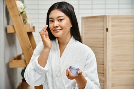 cheerful asian woman with acne applying cream on face and smiling while looking at camera at homeの写真素材