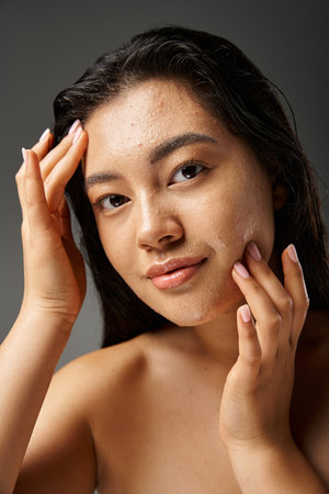 young asian woman with brunette hair toughing wet face with acne and looking at camera on greyの写真素材