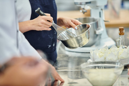 cropped view of young woman with apron whisking ingredients next to chief cook, confectionery