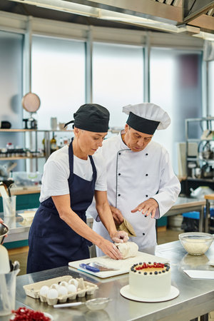 attractive mature chef in apron and toque working with dough next to her chief cook, confectionary