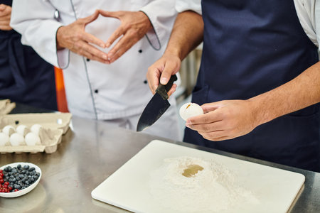 cropped view of african american chef in blue apron breaking egg with knife next to his colleaguesの写真素材