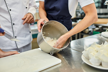 cropped view of mature female chef in blue apron working with dough next to her chief cook