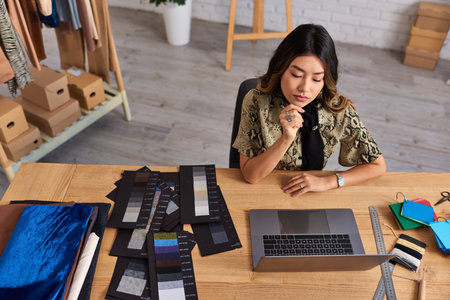 high angle view of serious asian fashion designer working at laptop near color samples in atelier