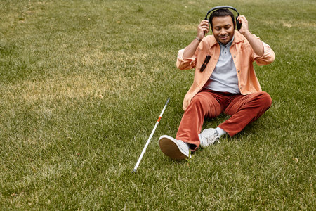 merry indian blind man in orange jacket sitting on grass with headphones and walking stick