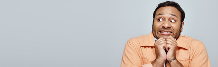 scared indian man in bright orange jacket posing and looking away on gray backdrop, banner