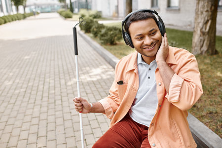 cheerful disabled indian man in casual outfit with headphones and walking stick enjoying musicの写真素材