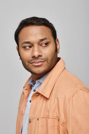 joyous indian man in vivid orange jacket smiling happily and looking away on gray backdrop