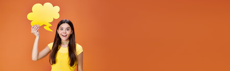 joyful teenager holding yellow thought bubble and looking at camera on orange backdrop, bannerの写真素材
