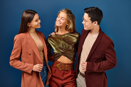 jolly adorable multiracial friends in fashionable attires smiling happily while on blue backdrop