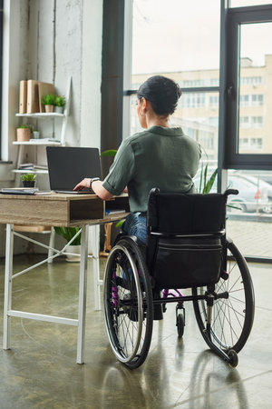 dedicated brunette disabled woman in casual attire sitting in wheelchair and working hard in officeの写真素材