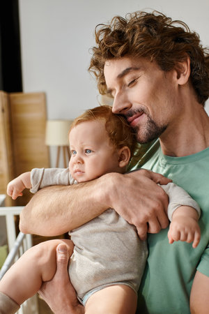curly-haired and cheerful father holding in arms his infant son in cozy bedroom at home, fatherhood