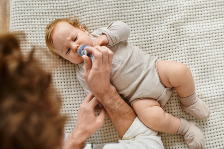 top view of single father giving pacifier to his infant baby son on the bed, fatherhood and love