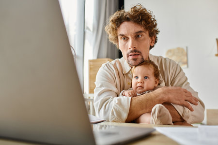 busy single father holding his cute infant baby in hands while working from home, work-life balanceの写真素材
