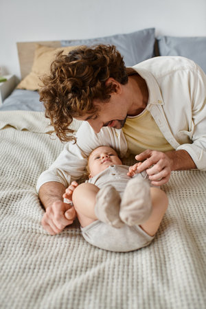 joyful father with curly hair and beard looking at his infant baby son on bed, precious moments