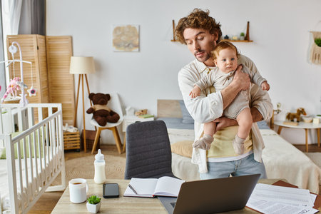 busy single father holding infant son and looking at papers near gadgets and baby bottle on deskの写真素材