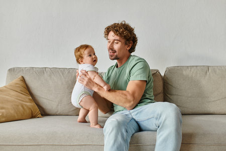 father holding in arms his barefoot infant baby son and spending quality time in living room