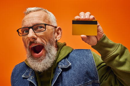 cheerful handsome mature male model in vibrant attire holding credit card on orange background