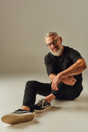attractive jolly mature man with glasses in black t shirt sitting on floor and smiling at cameraの写真素材