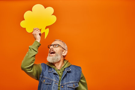 joyous handsome mature man in denim vest holding thought bubble above his head and looking at it