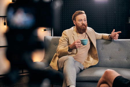 handsome bearded man with red hair in elegant clothes sitting next to his interviewer in studio