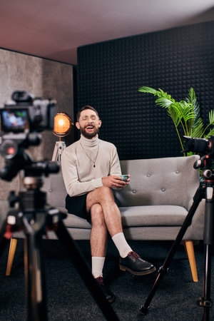 cheerful bearded man in elegant attire with glasses holding coffee cup during interview in studio