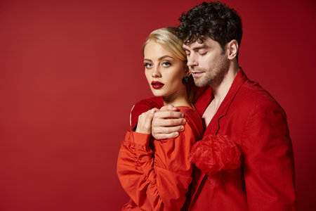 handsome man embracing blonde woman in elegant attire on red background, fashion