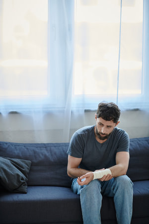 depressed man with bandage on arm after attempting suicide sitting on sofa, mental health awareness